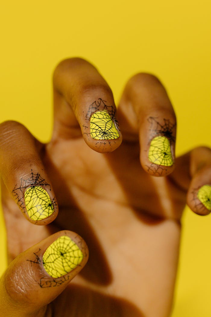 Bright yellow nails with black spider web design on fingers in a close-up shot on yellow backdrop.